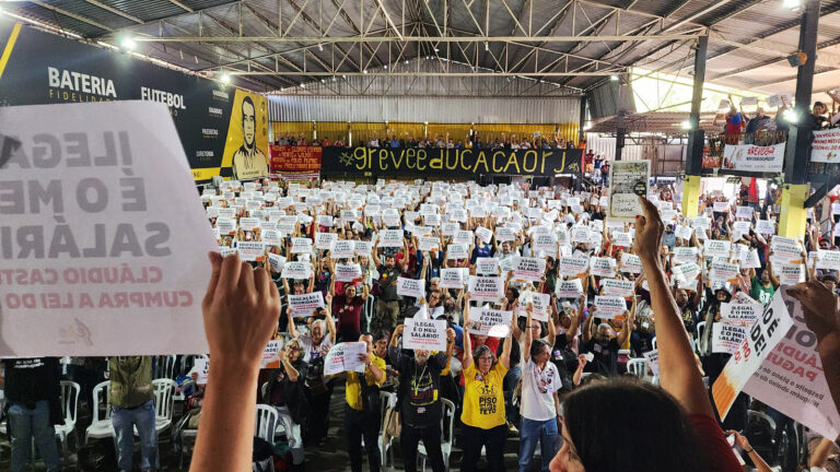 Imagem mostra assembleia dos professores no Rio de Janeiro em uma quadra. Os professores seguram cartazes reivindicando melhores salários.